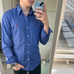 Blå Ralph Lauren skjorta  - Snygg blå skjorta från Ralph Lauren i slim fit med klassisk krage och vita knappar. Broderad röd logga på bröstet. Tillverkad i bomullstyg med beach twill-känsla. Perfekt för dig som gillar stilrena och tidlösa plagg.