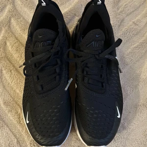 Nike Air Max 270 svarta sneakers - Svarta Nike Air Max 270 sneakers med vit sula och tydlig Air Max-bubbla i hälen. Ovandelen är i mesh-liknande syntetmaterial med snörning och små vita Nike-loggor på sidan och tån. Snygg och sportig design som passar perfekt till streetwear.