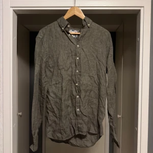 Olivgrön linneskjorta med knappar - Snygg olivgrön skjorta i linne med klassisk krage och vita knappar hela vägen. Skjortan har lång ärm och en avslappnad passform, perfekt för en stilren och chill look. Materialet är svalt och passar bra till varmare dagar.