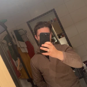 Brun half zip tröja Tommy Hilfiger - Säljer en brun långärmad tröja från Tommy Hilfiger med half zip och liten logga på bröstet. Tröjan har ribbade muddar och är i mjukt material, perfekt för höst och vinter. Snygg och enkel att matcha med jeans eller chinos.