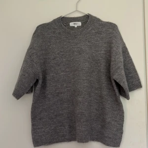 Grå stickad tröja från Gina Tricot - Mysig grå stickad tröja från Gina Tricot i ullblandning. Tröjan har korta ärmar, rund halsringning och en relaxed fit som passar perfekt till lager-på-lager. Enkel och stilren design som funkar till allt.