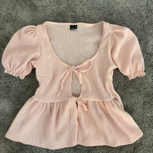 Ljusrosa blus med knyt - Supersöt ljusrosa blus från Gina Tricot med puffiga korta ärmar och två knyt framtill. Blusen har en v-ringad hals och är tillverkad i ett mjukt, strukturerat bomullstyg. 