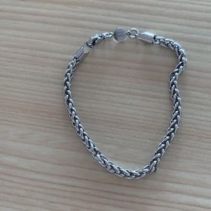 Silverfärgat kedjearmband från Safira - Stilrent armband i silverfärg från Safira med snyggt flätad kedja och diskret rund bricka med S-logga vid låset. Perfekt för dig som gillar minimalistiska accessoarer med lite edge. Lätt att matcha med andra smycken och har ett smidigt karbinlås.