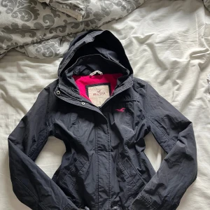 Svart vindjacka från Hollister S - Svart vindjacka från Hollister med huva och dragkedja. Insidan är fodrad med ceriserosa fleece för extra värme. Jackan har två fickor framtill och broderad Hollister-logga på bröstet och ryggen. Perfekt för blåsiga dagar.