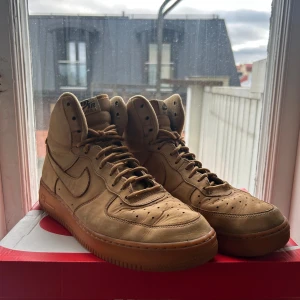 Nike Air Force 1 High sneakers - Säljer ett par Nike Air Force 1 High i färgen wheat/beige/khaki/brun med klassisk gum-sole. Skorna har högt skaft, snörning och är tillverkade i mocka med ikonisk swoosh på sidan. Perfekta för dig som vill ha en clean och stilren sneaker med streetkänsla.