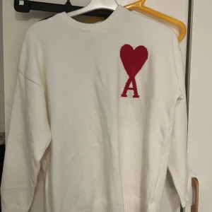 Vit stickad tröja Ami Paris hjärta - Vit stickad tröja från Ami Paris med röd hjärt- och A-logga på bröstet. Tröjan har rund hals, ribbade muddar och lång ärm. Perfekt för dig som gillar stilrena plagg med en twist.