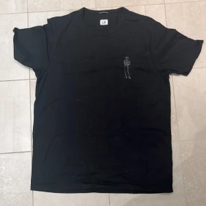 Svart t-shirt från C.P. Company - Svart t-shirt från C.P. Company i 100% merceriserad bomull. Framtill finns ett litet tryck av en figur och på ryggen ett större tryck med samma motiv samt loggan. Klassisk rund hals och korta ärmar. Snygg och stilren design med coolt print.
