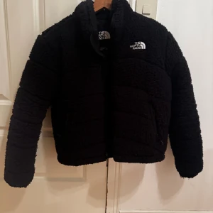 Svart teddyjacka från The North Face - Mysig svart teddyjacka från The North Face med hög krage och dragkedja. Jackan är croppad och har en oversized passform, perfekt för kalla dagar. Tillverkad i fluffigt teddy-material och pryds av logga på bröstet. Snygg och trendig att bära till både jeans och tights.