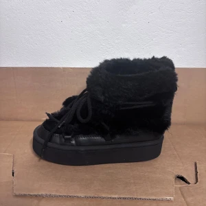 Svarta fluffiga boots med platåsula - Säljer ett par svarta boots med fluffig pälsimitation och snörning framtill. Skorna har en hög platåsula och är helsvarta, vilket ger en cool och mysig vibe. Perfekta för att hålla värmen och sticka ut i vintermörkret.