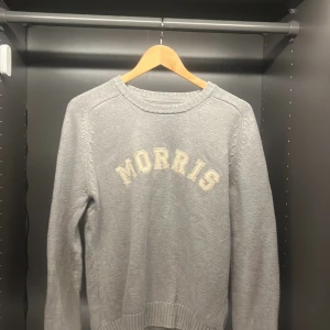 Grå stickad tröja från Morris - Snygg grå stickad tröja från Morris med broderad logga i vitt framtill. Tröjan har rund halsringning, ribbade muddar och lång ärm. Perfekt för dig som gillar klassisk och stilren look med en sportig touch med väldigt snygga detaljer.