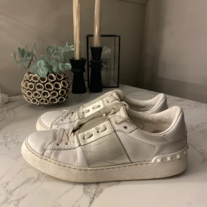 Valentino Garavani vit/silver sneakers med nitar - Snygga vita sneakers från Valentino Garavani med silvriga detaljer på sidorna och ikoniska nitar runt hälen. Skorna är i skinn och har snörning framtill. Perfekta för dig som vill ha en lyxig och trendig look.