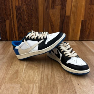 Nike Air Jordan 1 Low x Travis Scott  -  Ingen låda.Säljer ett par Nike Air Jordan 1 Low Travis Scott med svart, vit och blå färg. Perfekta för dig som gillar unika sneakers och streetwear.  