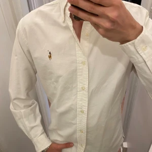 Vit Skjorta från Polo Ralph Lauren (Slim Fit) - Vit skjorta från Ralph Lauren med klassisk button-down krage och broderad logga på bröstet. Skjortan har lång ärm, knappar framtill och är gjord i mjuk bomull. Perfekt för dig som gillar stilrena och tidlösa plagg med snygga detaljer. Storlek Medium. Snabb Leverans!