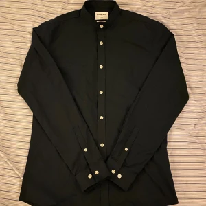 Svart skjorta från Lindbergh - Stilren svart skjorta från Lindbergh med klassisk ståkrage och vita knappar längs hela framsidan. Skjortan är långärmad och tillverkad i mjuk bomull, perfekt för dig som gillar minimalistisk och clean look.