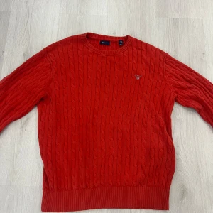 Röd kabelstickad tröja från GANT - Snygg röd kabelstickad tröja från GANT med rund hals och diskret logga broderad på bröstet. Tröjan har ribbade muddar vid hals, ärmslut och nederkant. Perfekt för dig som gillar klassisk stil med en färgklick.