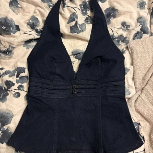 Mörkblå denim halterneck korsett - Snygg mörkblå korsettopp i denim med halterneck och markerad midja med dubbla dekorativa band framtill. Toppen har djup v-ringning och dragkedja i ryggen. Perfekt för dig som vill ha en edgy och trendig look.
