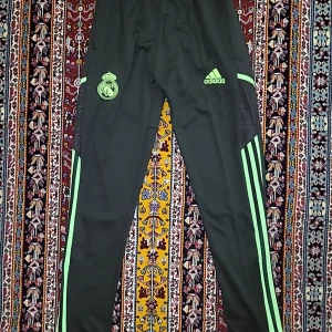 Adidas Real Madrid träningsbyxor svart - Svarta träningsbyxor från Adidas med Real Madrid-logga och gröna detaljer. Byxorna har smal passform, gröna ränder längs sidorna och är tillverkade i ett lätt syntetmaterial. Perfekta för fotbollsträning eller chill. Elastisk midja och dragkedja vid benslut.