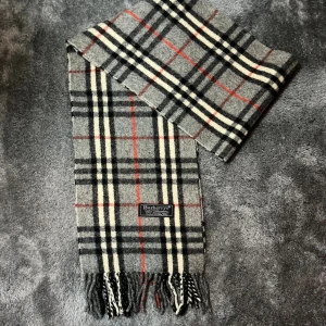 Rutig halsduk från Burberrys i ull - Klassisk rutig halsduk från Burberrys i 100% lammull. Färgkombinationen är grå, svart, vit och röd med fransar i ändarna. Den ikoniska rutiga designen gör den till ett stilrent statement till vinterns outfits.❄️