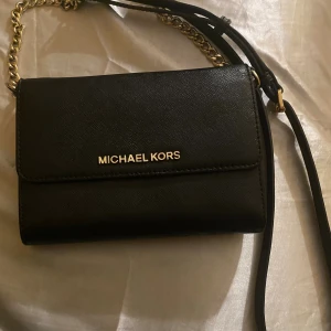 Svart axelväska från Michael Kors - Snygg svart axelväska från Michael Kors i skinn med guldfärgade metalldetaljer och kedja på axelremmen. Väskan har stilren design, flera fack och kortplatser inuti samt logga i guld framtill. Perfekt för dig som gillar klassisk och lyxig stil. I Saffiano leather. 