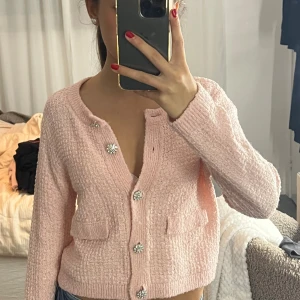 Ljusrosa kofta med glittriga knappar - Superfin ljusrosa kofta med glittriga blomformade knappar och två små fickor framtill. Koftan är stickad och har en croppad passform med långa ärmar. Perfekt för att ge en söt touch till din outfit!