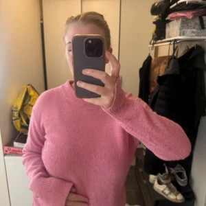 Rosa stickad tröja med rund hals - Mysig rosa stickad tröja med rund halsringning och ribbade muddar vid ärmslut och nederkant. Tröjan har en relaxed passform och långa ärmar, perfekt för lager-på-lager under kyliga dagar. Enkel och stilren design som är lätt att matcha. Den är nopprig