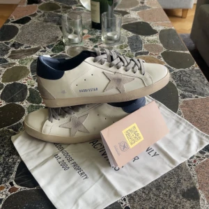Golden Goose Superstar sneakers, vit/grå - Golden Goose Superstar sneakers i vitt skinn med grå mockadetaljer och stjärna på sidan. Mörkblå häl, grå snörning och beige sula. Klassisk låg modell med rund tå och platt sula. Snygg italiensk design, perfekt för dig som gillar streetstyle.