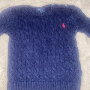 Marinblå kabelstickad tröja Polo Ralph Lauren - Kabelstickad marinblå tröja från Polo Ralph Lauren med rund halsringning, långa ärmar och knappar på axeln. Tröjan har en röd broderad logga på bröstet och är gjord i mjuk bomull. Perfekt för dig som gillar klassisk preppy stil. Barn storlek