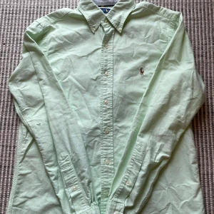Ljusgrön skjorta från Ralph Lauren - Snygg ljusgrön skjorta från Ralph Lauren med klassisk button-down krage och broderad logga på bröstet. Skjortan har långa ärmar, knäppning framtill och är tillverkad i mjuk bomull. Perfekt för dig som gillar stilrena och tidlösa plagg.