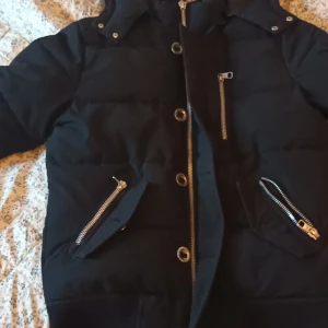 Svart pufferjacka med silverdetaljer - Svart pufferjacka från TD med huva, stora silverfärgade dragkedjor och knappar framtill. Jackan har en broderad TD-logga på ärmen och flera fickor med dragkedja. Snygg och varm modell med normal passform, perfekt för kalla dagar.