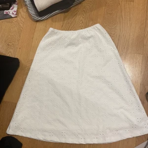 Vit broderad långkjol i bomull - Superfin vit långkjol i bomull med broderade detaljer och små hålmönster. Kjolen har en lätt A-linjeform och resår i midjan för skön passform. Perfekt för varma dagar och ger en fräsch och romantisk vibe.