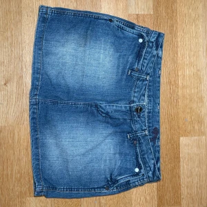 Blå jeanskjol från Wrangler W26 - Snygg blå lågmidjad mini jeanskjol från Wrangler i klassisk femficksmodell med bälteshällor och detaljerade bakfickor med logopatch. Kjolen är tillverkad i 100% bomull och har en kort, rak passform som ger en cool och avslappnad vibe.