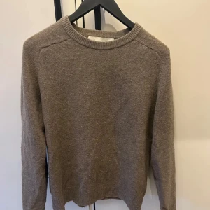 Beige stickad tröja med rund hals - En enkel och stilren beige stickad tröja med rund halsringning och långa ärmar. Perfekt att slänga på sig när det är kyligt ute. Tröjan har ribbade muddar vid hals, ärmslut och nederkant för en klassisk look.