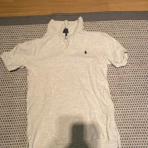 Vit pikétröja från Polo Ralph Lauren - Säljer en klassisk vit pikétröja från Polo Ralph Lauren med korta ärmar och den ikoniska loggan broderad på bröstet. Tröjan har krage och knappar framtill, perfekt för en clean och stilren look.