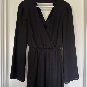 Svart klänning - Svart kort klänning med lång ärm och markerad midja. Klänningen har v-ringning framtill med en knapp. Knäppning i nacken. 