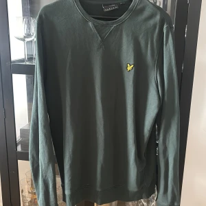 Mörkgrön sweatshirt från Lyle & Scott - Mörkgrön sweatshirt från Lyle & Scott med klassisk rund halsringning och gul logga på bröstet. Tröjan har långa ärmar och ribbade muddar vid ärmslut och nederkant. Perfekt för en chill och stilren look.