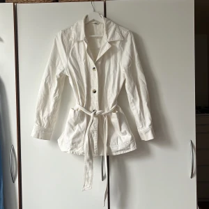 Vit jacka med bälte från & Other Stories - En stilren, fin, vit jacka från & Other Stories i 100% bomull. Jackan har klassisk krage, knappar framtill, två stora fickor, långa ärmar med knappdetalj och ett matchande bälte i midjan. 