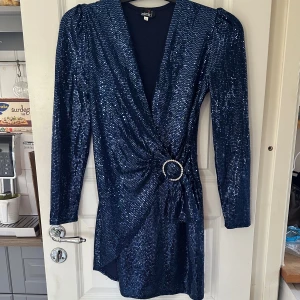 Marinblå glittrig klänning från Zack - Snygg marinblå kortklänning från Zack London med glittriga paljetter över hela klänningen. Djup v-ringning, markerade axlar och lång ärm. Snygg detalj med glittrande bälte i midjan. Perfekt för dig som vill sticka ut med en glamorös look. Använd 1 gång på nyår. 