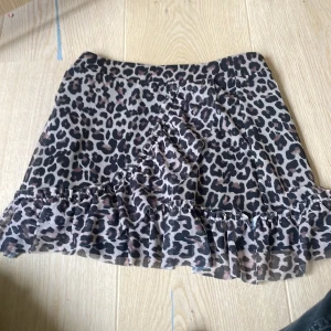 Leopardmönstrad volangkjol från Lindex - Säljer en kort kjol från Lindex med leopardmönster i svart och beige. Kjolen har volangdetalj längs nederkanten och omlottkänsla framtill. Perfekt för dig som vill sticka ut med ett djurmönstrat plagg.