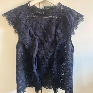 Mörkblå spetsblus med blommönster - Säljer en mörkblå blus i spets med blommigt mönster och volanger. Blusen är ärmlös med  spetskanter och har en liten öppning bak i nacken. Storlek M men passar mig som är en S!