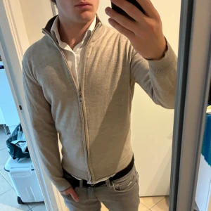 Beige stickad kofta med dragkedja - Snygg beige stickad kofta från okänt märke, hel dragkedja framtill och ribbad krage. Modellen är långärmad och har en clean, stilren look som funkar till skjorta eller t-shirt. Perfekt för dig som gillar enkel och klassisk stil. Storlek M men sitter som XS. Ett par lagningar på insidan av armarna, inget som syns utanpå.