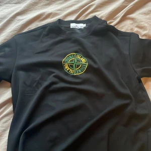 Svart Stone Island T-shirt - Svart sweatshirt från Stone Island med klassisk kompasslogga broderad på bröstet och avtagbar patch på ärmen. Tröjan har rund hals och är tillverkad i mjuk bomull. Perfekt för dig som gillar streetwear och vill ha en stilren look.