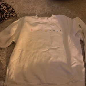 Vit Nike Golf sweatshirt - Säljer en clean vit sweatshirt från Nike Golf med broderad logga i flera färger på bröstet. Tröjan har rund halsringning och långa ärmar, perfekt för en sportig och avslappnad stil. Mjuk och skön, passar dig som gillar enkel men snygg design.