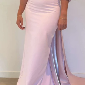 Ljusrosa långklänning med pärlor - Superfin ljusrosa långklänning med offshoulder och stor rosett baktill. Klänningen har pärldetaljer på överdelen och en elegant slits framtill. Materialet är glansigt och faller snyggt längs kroppen. Endast använd en gång!