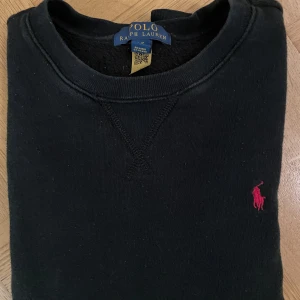 Svart sweatshirt från Polo Ralph Lauren - Svart sweatshirt från Polo Ralph Lauren med klassisk rund hals och broderad rosa logga på bröstet. Tröjan har långa ärmar och ribbade muddar vid ärmslut och nederkant. Perfekt för en chill och stilren look.