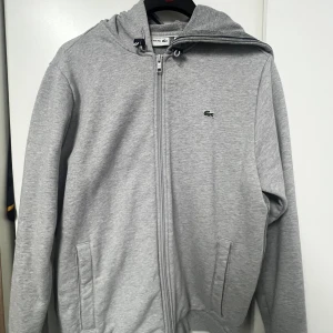 Grå hoodie från Lacoste med dragkedja - Säljer en stilren grå hoodie från Lacoste med hel dragkedja och klassisk krokodillogga på bröstet. Tröjan har huva med snörning, två fickor framtill och är tillverkad i mjuk bomullsmix. Perfekt för chill dagar eller när du vill ha en clean look. Storlek L