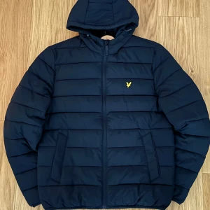 Svart pufferjacka från Lyle & Scott - Snygg Svart pufferjacka från Lyle & Scott med huva och dragkedja. Jackan har det klassiska gula logobroderiet på bröstet och två stora fickor framtill. Perfekt för kyliga dagar och riktigt skön att bära.