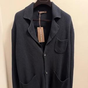 Oscar Jacobson cardigan  - Snygg mörkblå stickad cardigan från Oscar Jacobson med krage, tre framfickor och knappar. Cardiganen har en grovstickad struktur och avslappnad passform, perfekt för dig som gillar stilren look