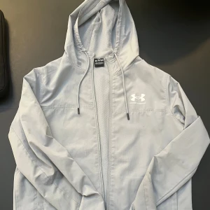 Ljusgrå vindjacka från Under Armour - Snygg ljusgrå vindjacka från Under Armour med huva och snörning. Säljer den då den inte riktigt passar min stil längre. Tveka inte att kontakta för mer bilder om det behövs😊