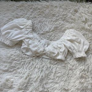 Vit croppad blus med puffärmar - Superfin vit croppad blus med puffiga korta ärmar och smockdetaljer. Toppen har snörning framtill och är gjord i mjuk bomull med lite stretch. Perfekt för en trendig och luftig look under varma dagar.