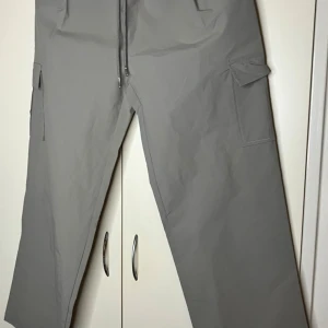 Grå cargopants med snörning - Säljer ett par grå cargopants med bred passform och praktiska fickor på sidorna. Byxorna har snörning i midjan för extra komfort och är tillverkade i ett mjukt syntetmaterial. Perfekta för en chill och avslappnad stil.